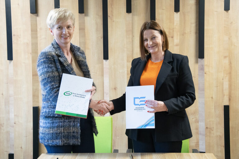 zdjęcie przedstawiające przedstawicielki Akademii Mazowieckiej w Płocku i Banku Spółdzielczego w Starej Białej na tle logotypów instytucji, trzymające teczki z podpisanym porozumieniem o współpracy, podające sobie ręce