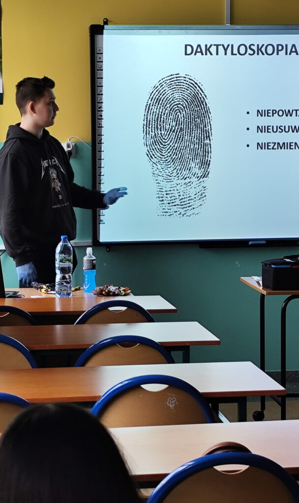 Student omawiający prezentację z kryminalistyki przed uczniami siedzącymi w ławkach szkolnych