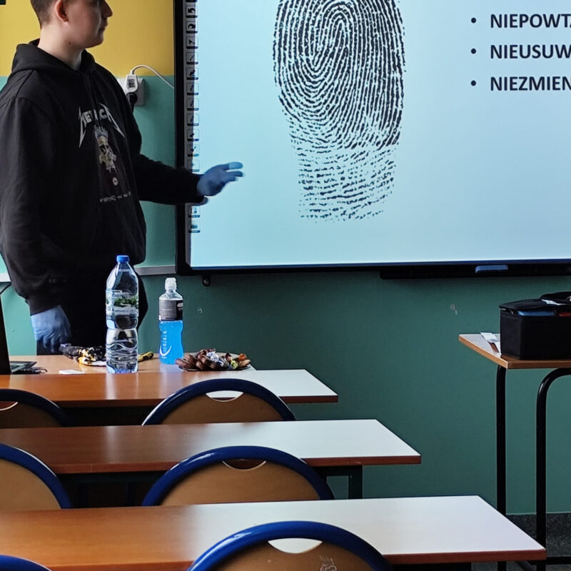 Student omawiający prezentację z kryminalistyki przed uczniami siedzącymi w ławkach szkolnych