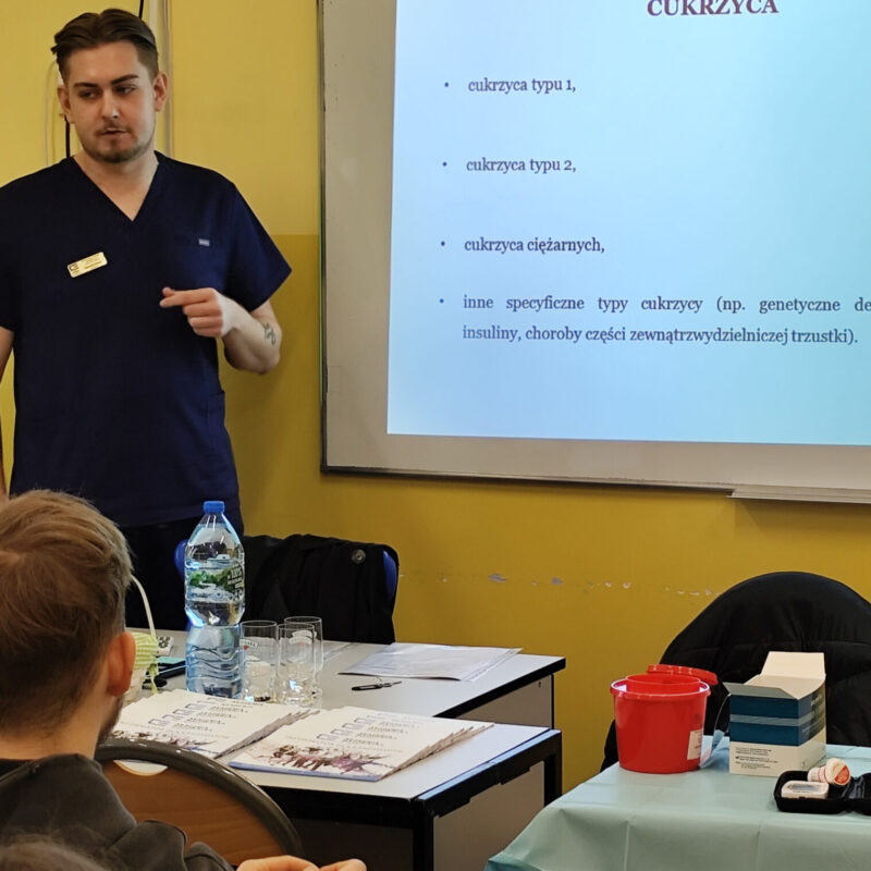 Student omawiający prezentację z podstaw oznaczania poziomu glikemii przed uczniami siedzącymi w ławkach szkolnych