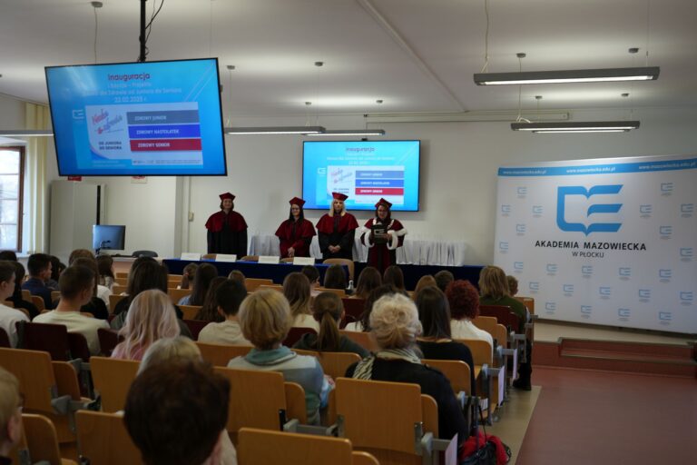 Realizatorzy projektu ze strony AMP stojący za stołem prezydialnym w strojach akademickich, przed uczestnikami, w trakcie oficjalnego otwarcia