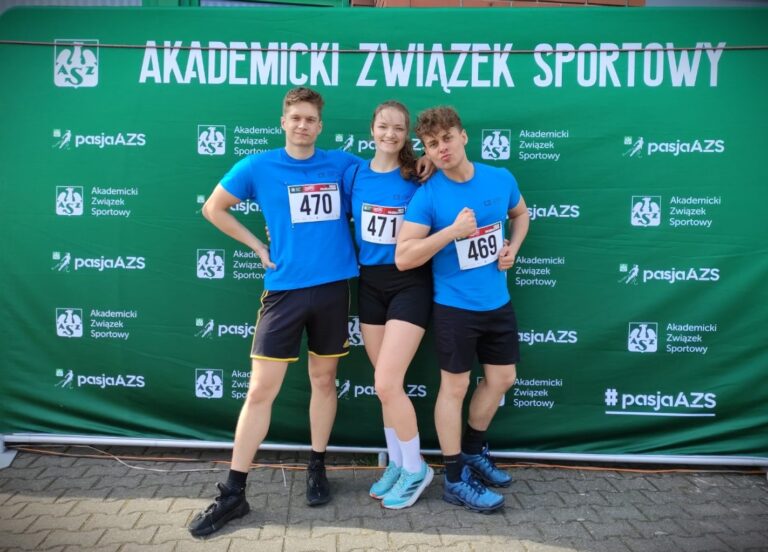 Studenci, dwóch mężczyzn i jedna kobieta, biorący udział w Akademickich Mistrzostwach Polski w biegach przełajowych w sportowych strojach pozujący na ściance Akademickiego Związku Sportowego