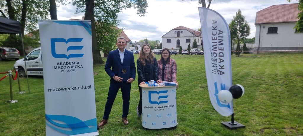 Przedstawiciele AMP Prodziekan i studentki stojący przy ladzie promocyjnej uczelni, przy rollupie, w plenerze