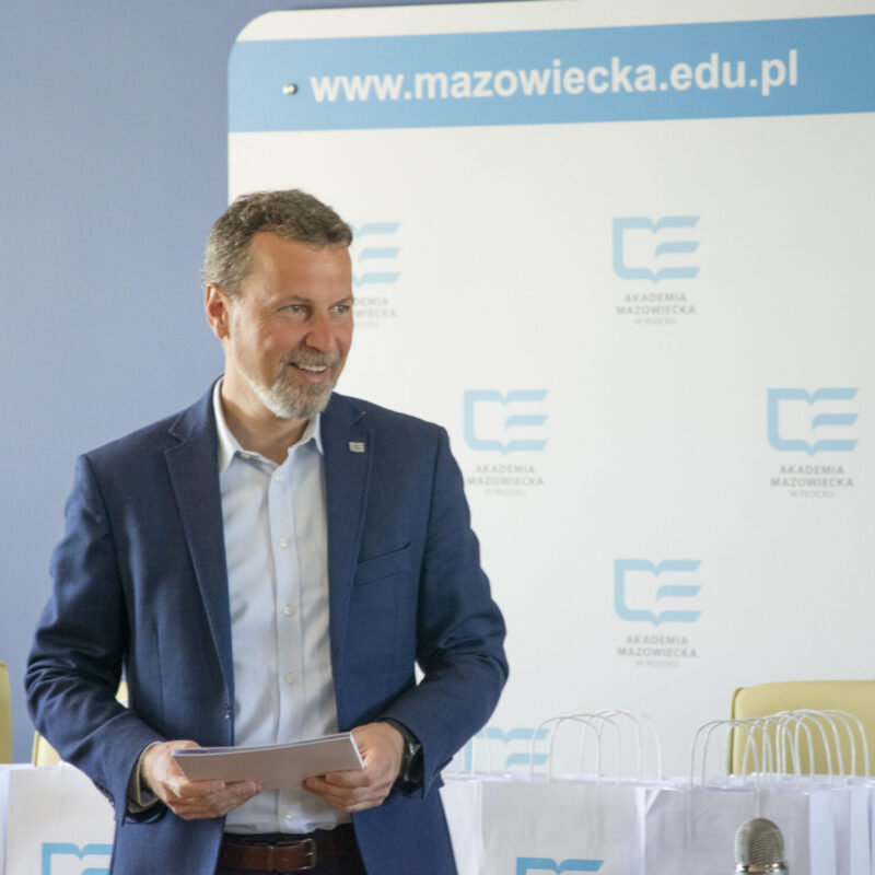 Uśmiechnięty Rektor podczas rozmowy ze studentami, w tle ścianka z logotypami uczelni