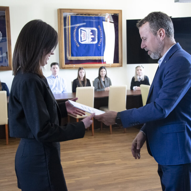 Rektor wręczający dyplom studentce, w tle siedzący przy stole studenci.
