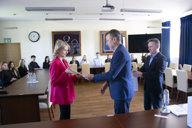 Rektor wręczający dyplom studentce, w towarzystwie prorektora, w tle siedzący przy stole studenci.