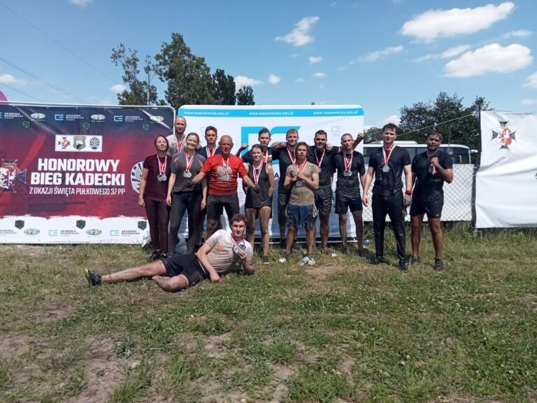 grupa studentów po zmaganiach biegowych wraz z profesorem Wiśniewskim pozująca do zdjęcia na tle ścianki Akademii Mazowieckiej w Płocku oraz ścianki Honorowego Biegu Kadeckiego