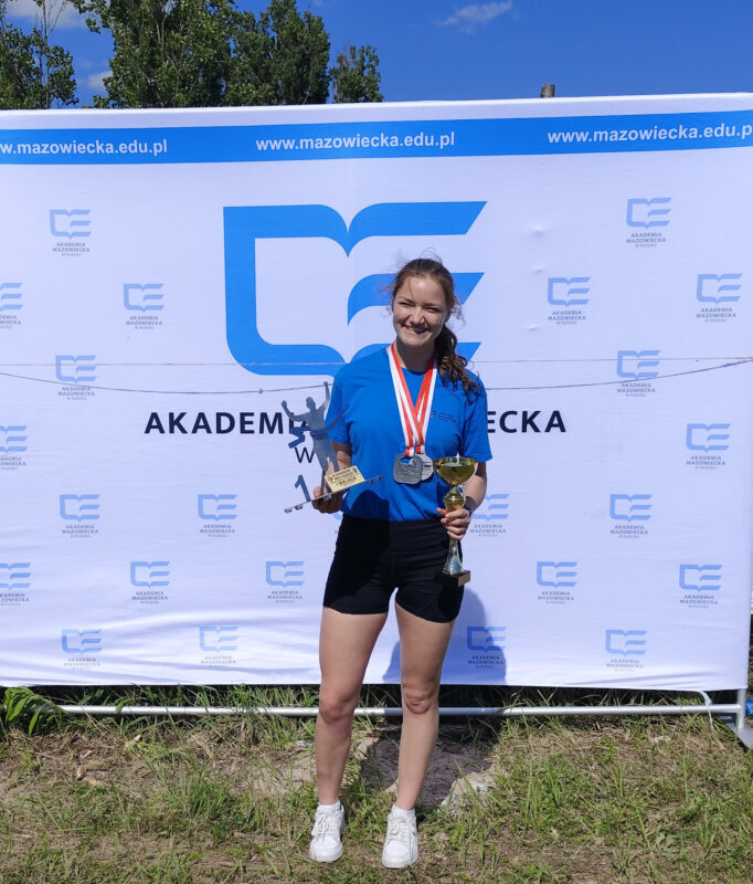 Zawodniczka reprezentująca Akademię Mazowiecką w Płocku pozująca do zdjęcia na tle ścianki z logo AMP z pucharami