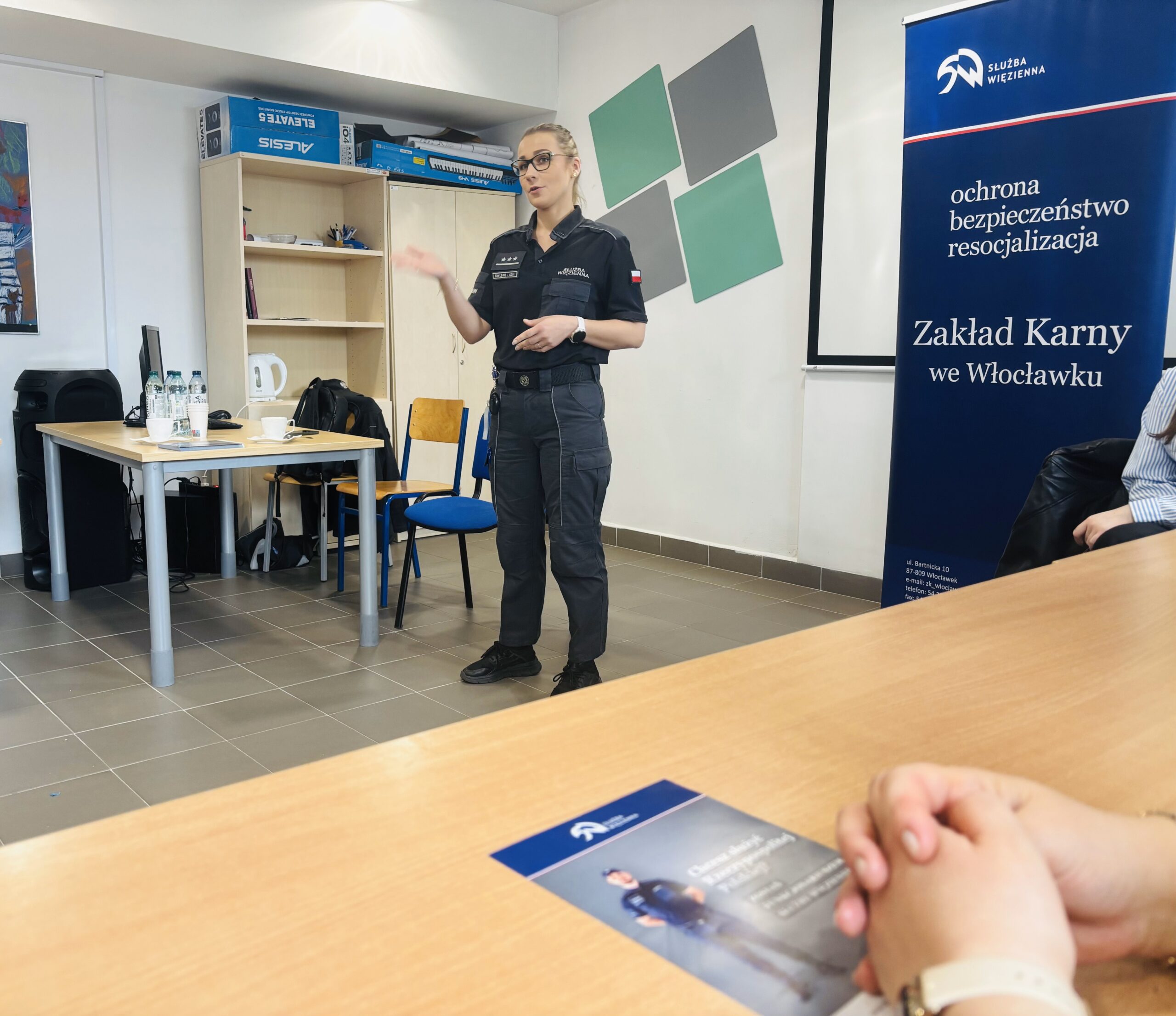 Kobieta w mundurze służby więziennej stojąca i opowiadająca w sali wykładowej AMP, w tle roll-up Zakładu Karnego we Włocławku.