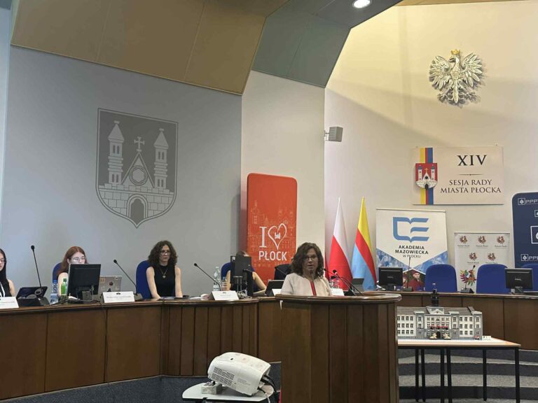 Kobieta przemawiająca w sali urzędu miasta, w tle siedzące osoby przy stole przezydialnym