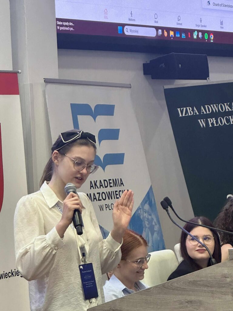 młoda osoba, kobieta, przemawiajaca do mikrofonuw Auli AMP w uniesiona lewą ręką, obok inne osoby siedzące przy stole przezydialnym