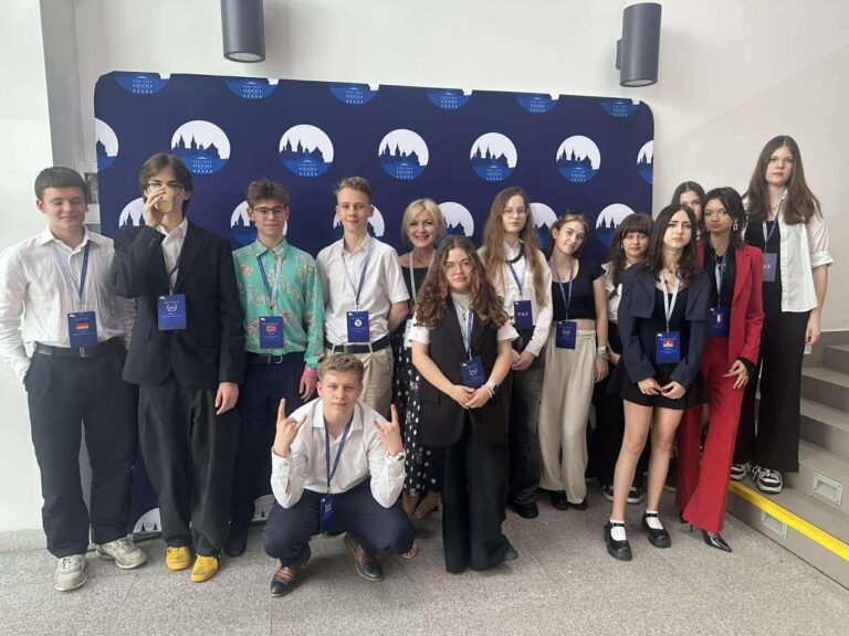 Grupa młodzieży, elegancko ubranych, z identyfikatorami na szyi, pozująca do zdjęcia na ściance 8 edycji konferencji Płock Model United Nations z opiekunką konferencji
