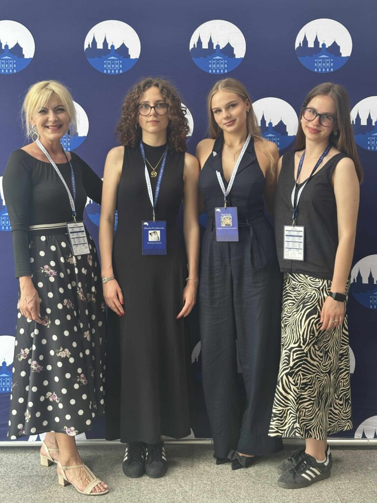 Grupa osób, elegancko ubranych, z identyfikatorami na szyi, pozująca do zdjęcia na ściance 8 edycji konferencji Płock Model United Nations z opiekunką konferencji