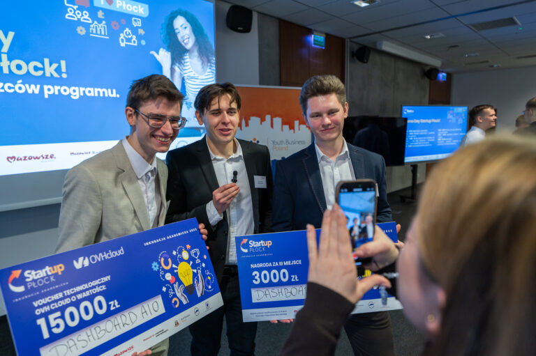 zdjęcie grupowe 3 mężczyzn, trzymajacych plansze z nagrodą, jeden z nich mówi do mikrofonu, są nagrywani na telefon. z tyłu ekran z prezentacją.