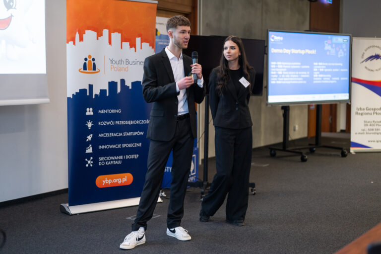 2 osoby- kobieta i mężczyzna stojący na tle rollupu, mężczyzna mówi do mikrofonu